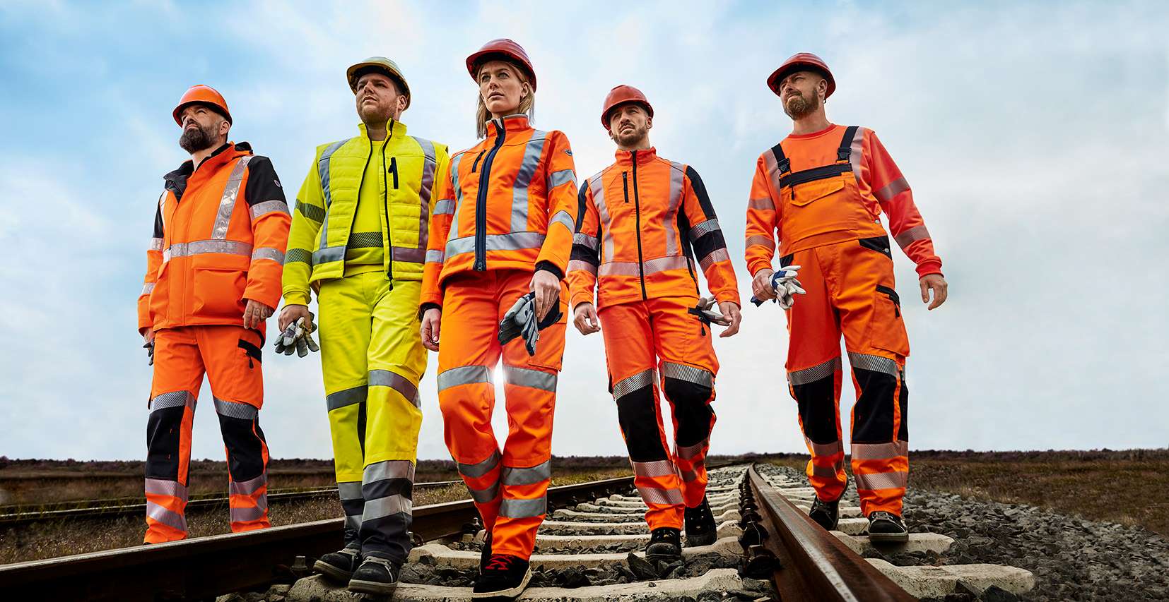 Vier Gleisarbeiter in orangefarbener und einer in neonfarbiger Warnschutzkleidung gehen auf einem Gleis.